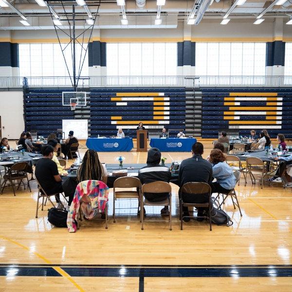 tables in a gymnasium with people seated around them; head table has blue drapes with GVSU logo. Bleachers in background are blue with large gold B and C showing